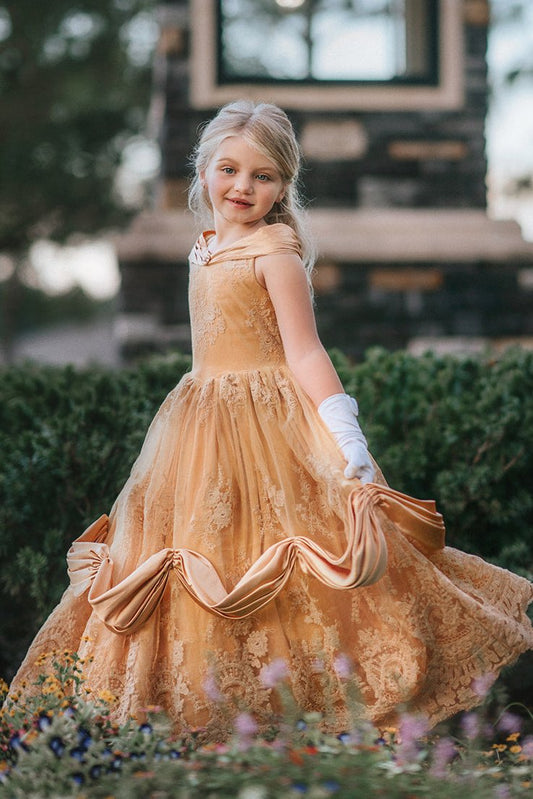 A young girl with blonde hair stands outdoors, wearing the Portrait Collection Red Rose Princess Gown, an intricately designed gold princess dress adorned with floral lace patterns. The satin off shoulder neckline is adorned with a gold jewel. 