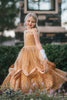 A young girl with blonde hair stands outdoors, wearing the Portrait Collection Red Rose Princess Gown, an intricately designed gold princess dress adorned with floral lace patterns. The satin off shoulder neckline is adorned with a gold jewel. 