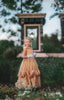 A young girl with blonde hair stands outdoors, wearing the Portrait Collection Red Rose Princess Gown, an intricately designed gold princess dress adorned with floral lace patterns. She gracefully holds up the hem of her gown and wears white gloves. Trees and a building with stone bricks form the backdrop, enhancing The Red Rose Princess ambiance.