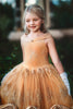 A young girl with blonde hair stands outdoors, wearing the Portrait Collection Red Rose Princess Gown, an intricately designed gold princess dress adorned with floral lace patterns. She gracefully holds up the hem of her gown and wears white gloves. Trees and a building with stone bricks form the backdrop, enhancing The Red Rose Princess ambiance.