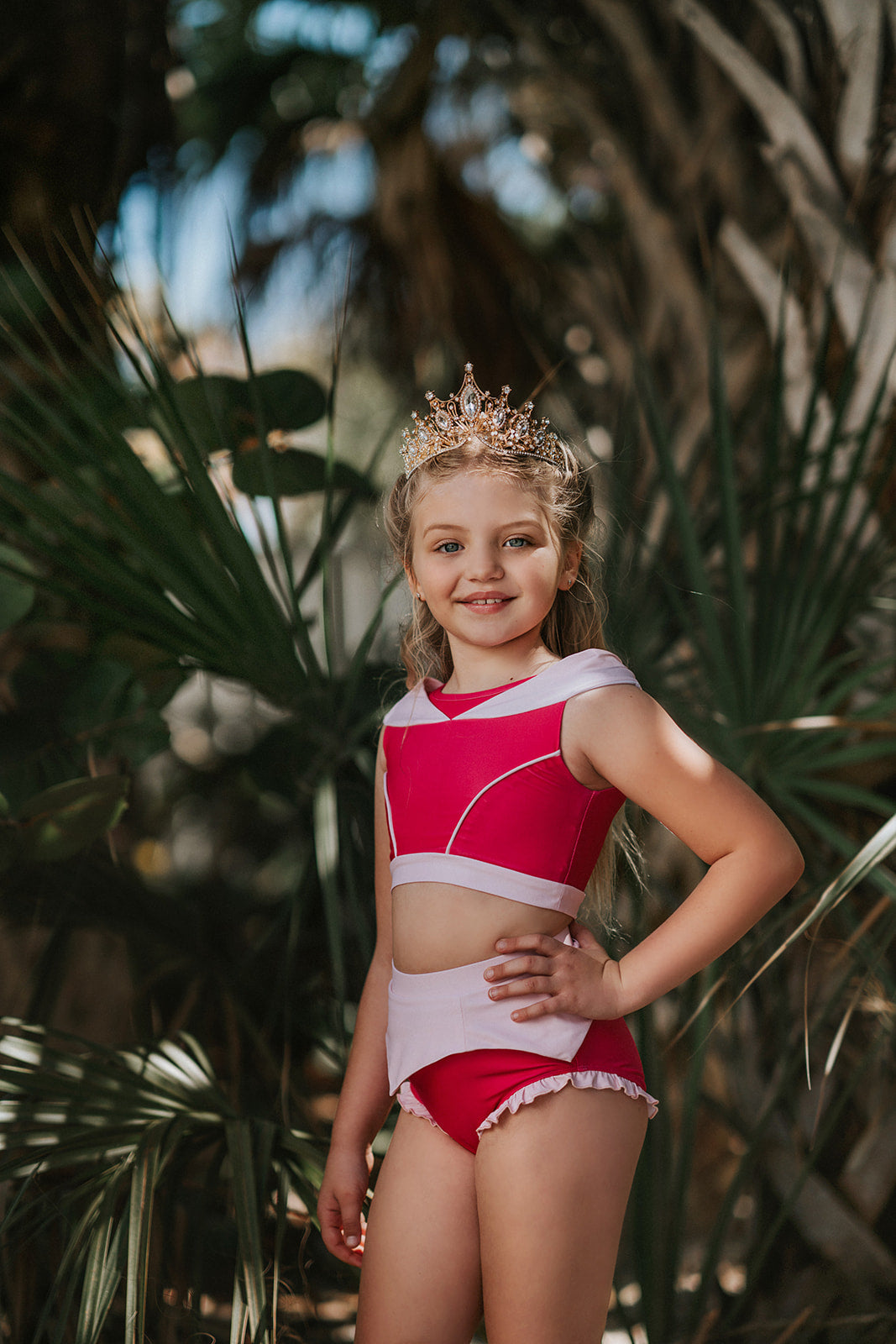 A young girl is wearing a pink 2 piece swimsuit.  The Sleeping Beauty swim set has a pink collar that resembles a ballgown.  The top has light pink piping and trim.  The modest bottoms have a pink overlay and ruffles on the legs. 