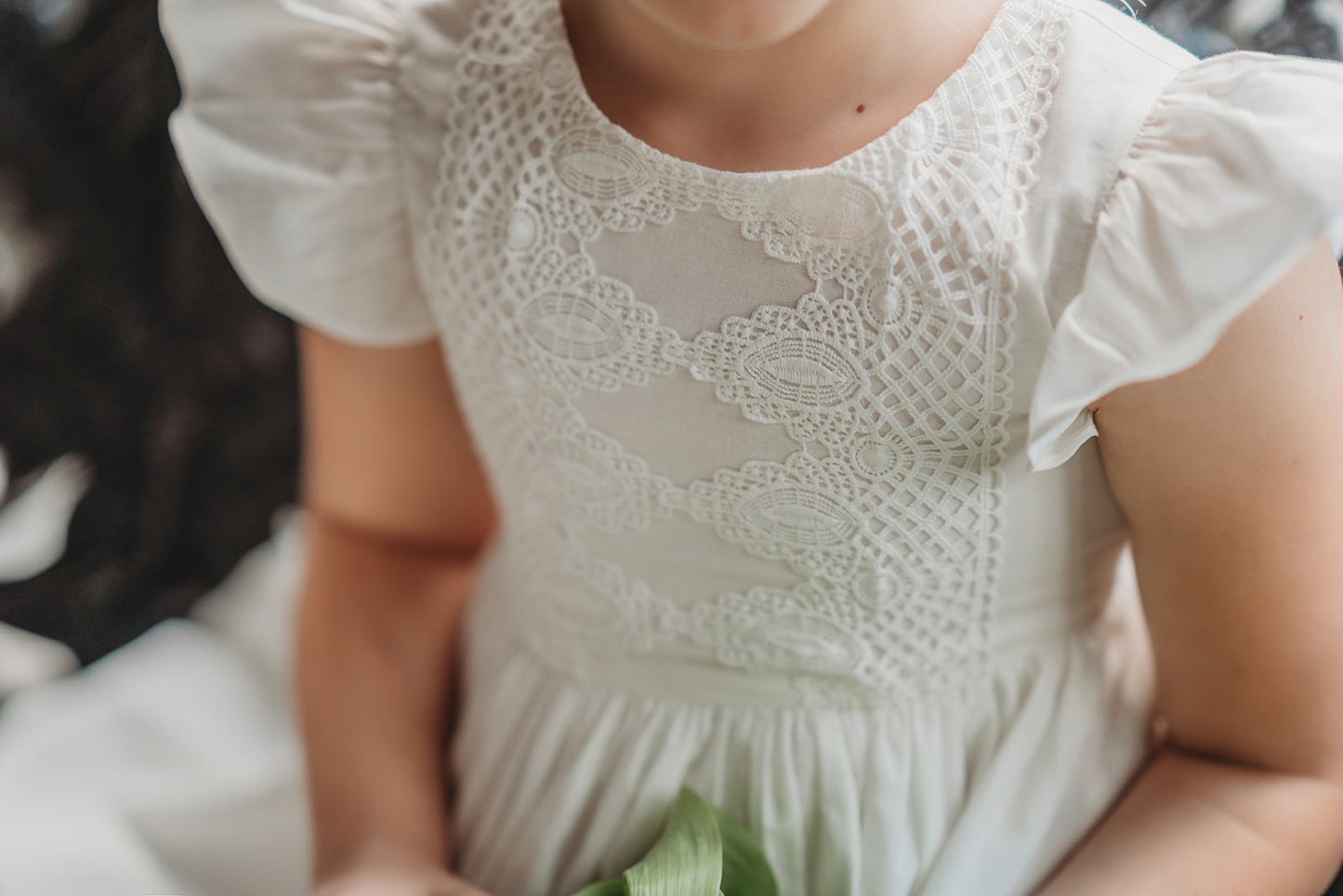 A young girl is wearing the Ethel Fancy Pinafore in Soft White with ruffled sleeves. There is a matching lace overlay on the bodice.  A pettiskirt can be added underneath the dress for fullness. 