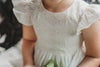 A young girl is wearing the Ethel Fancy Pinafore in Soft White with ruffled sleeves. There is a matching lace overlay on the bodice.  A pettiskirt can be added underneath the dress for fullness. 