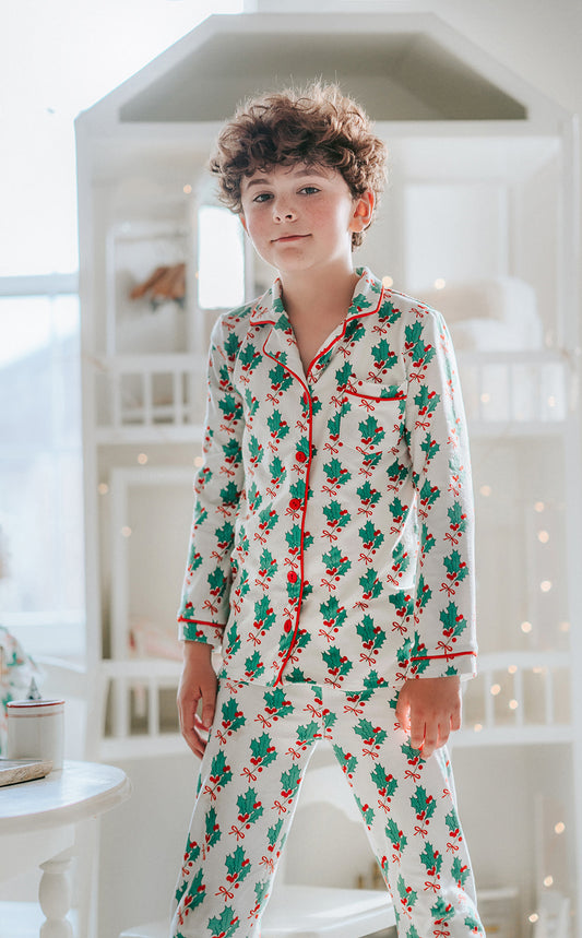 A young boy is wearing the classic Merry Mistletoe Lounge set.  This 2pc pajama has green mistletoe and red berries printed all over it.  The top has red piping and red buttons. 