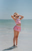 A young girl is wearing a 2 piece pink swim set. There is white collar with a pink bow at the neck.  Two lighter pink stripes are on the chest like ribbon tails.  The bottom is a skirt with bows light pink bows at the hem.