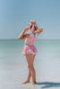 A young girl is wearing a 2 piece pink swim set. There is white collar with a pink bow at the neck.  Two lighter pink stripes are on the chest like ribbon tails.  The bottom is a skirt with bows light pink bows at the hem.