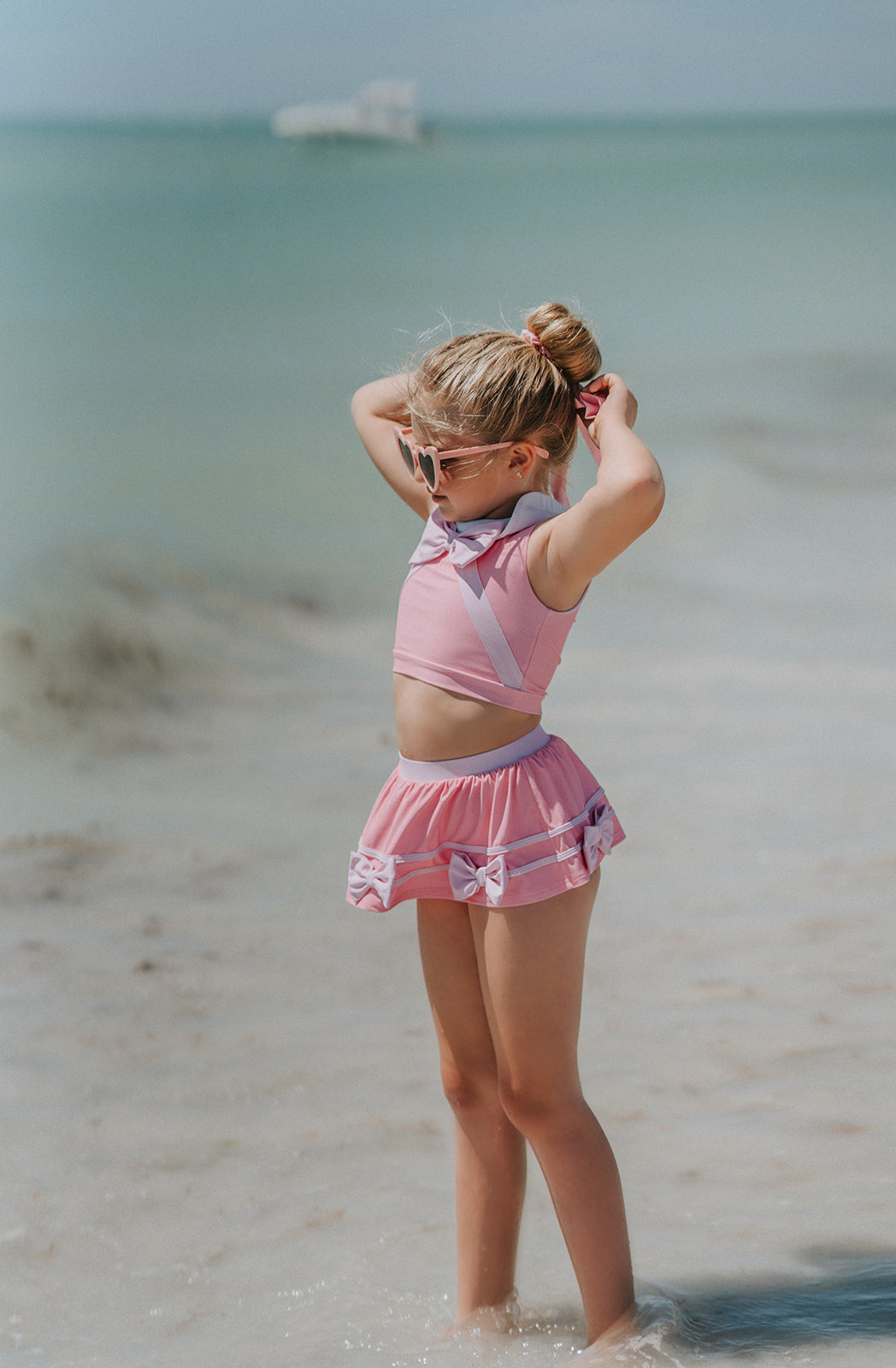 A young girl is wearing a 2 piece pink swim set. There is white collar with a pink bow at the neck.  Two lighter pink stripes are on the chest like ribbon tails.  The bottom is a skirt with bows light pink bows at the hem.