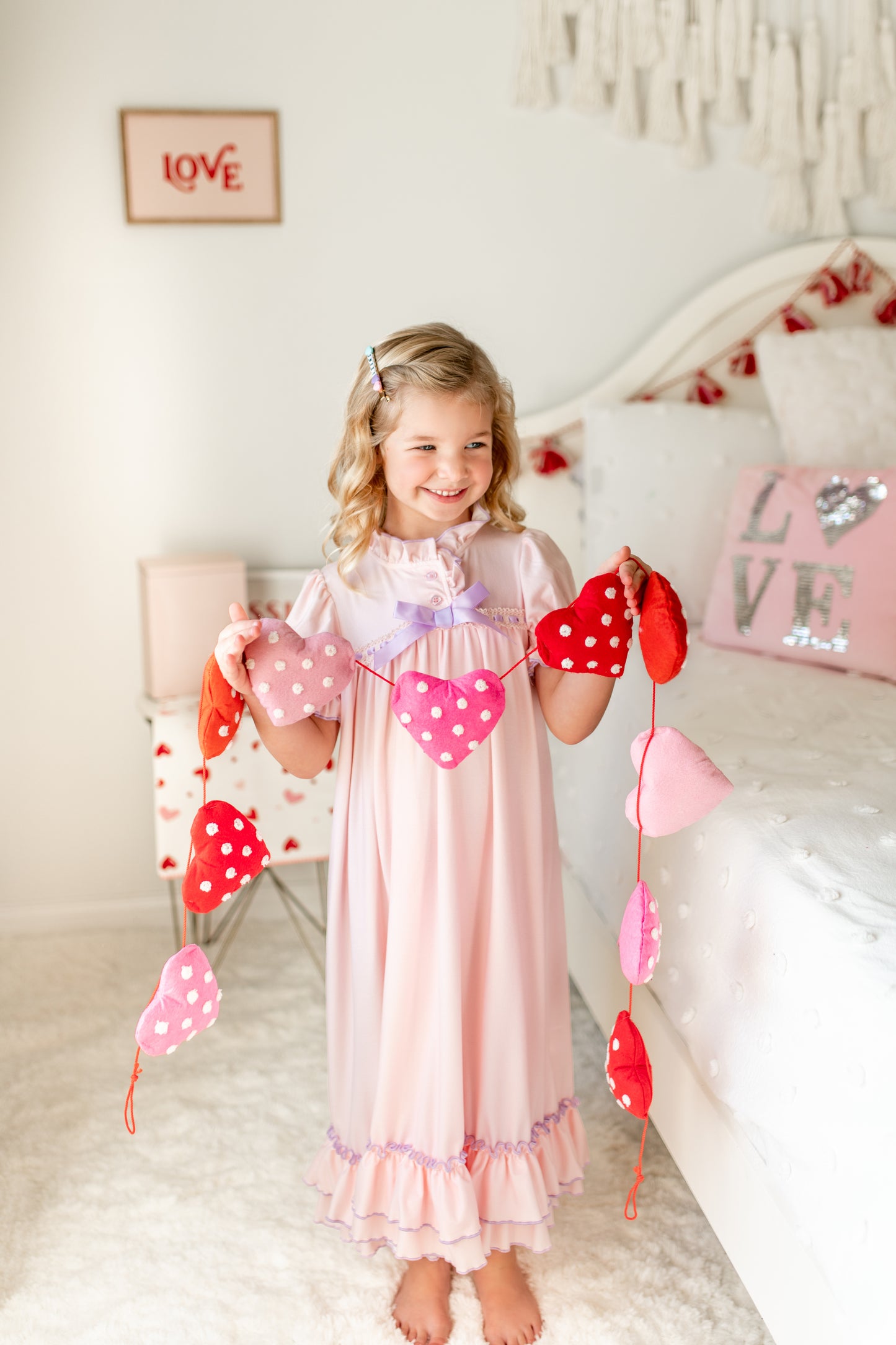 A young girl with curly blonde hair stands in a bright room wearing The Original Clara Nutcracker Gown in Blush, featuring purple ruffles and a bow. She has a smile on her face. Behind her, there's a white bed with decorative pillows and a sign on the wall that reads "LOVE." Her outfit looks like it's from Clara's children's sleepwear collection.