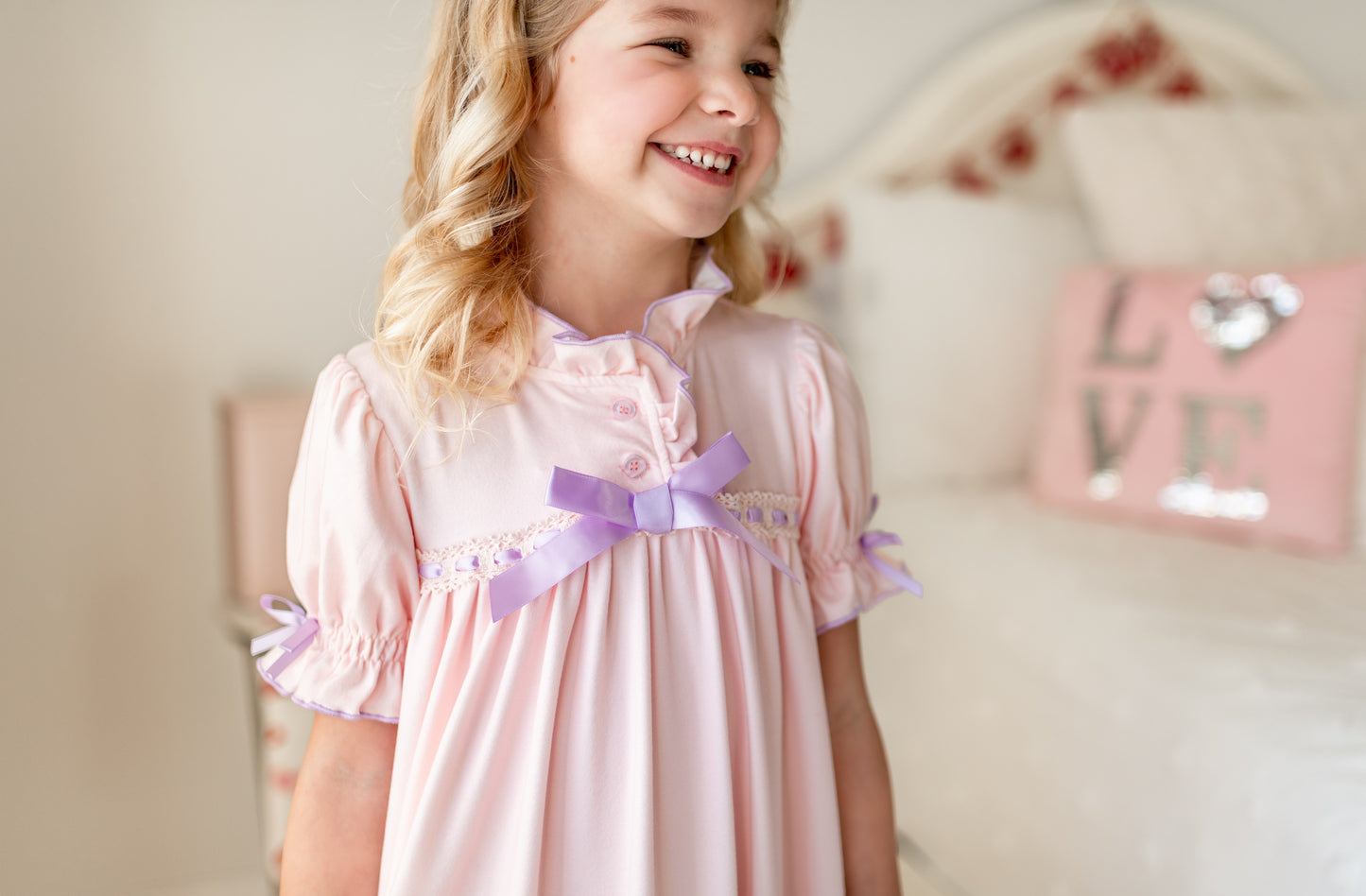 A young girl with curly blonde hair stands in a bright room wearing The Original Clara Nutcracker Gown in Blush, featuring purple ruffles and a bow. She has a smile on her face. Behind her, there's a white bed with decorative pillows and a sign on the wall that reads "LOVE." Her outfit looks like it's from Clara's children's sleepwear collection.