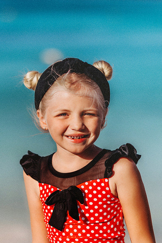 A young girl is wearing our Miss Mouse  2pc Swim set.  This 2pc swim has SPF 50.  The top is red with white polka dots.  There is a large black bow in the center and small ruffles on the shoulder.  The bottoms have black ruffles along the edge. 
