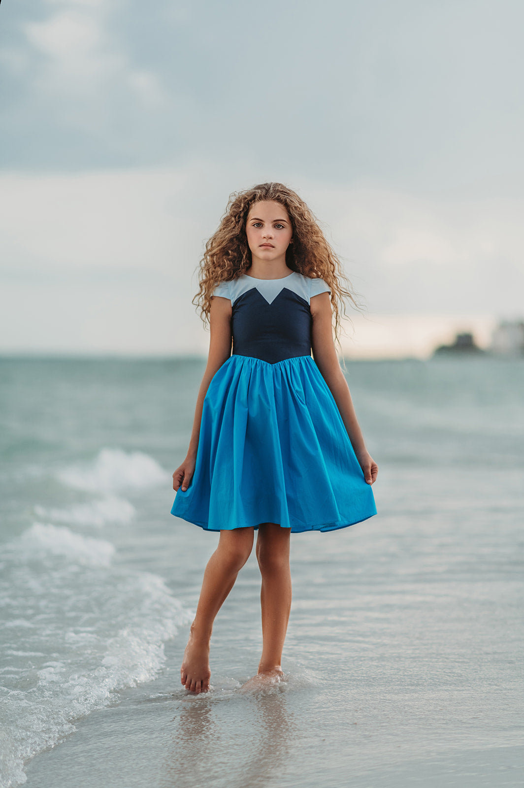 A young girl is wearing a Kiss the Girl Mermaid Dress from The Fairy Godmother Collection, featuring a navy bodice and light blue cap sleeves. The dress skirt is a bight cerulean blue. The material is woven cotton. 