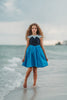 A young girl is wearing a Kiss the Girl Mermaid Dress from The Fairy Godmother Collection, featuring a navy bodice and light blue cap sleeves. The dress skirt is a bight cerulean blue. The material is woven cotton. 