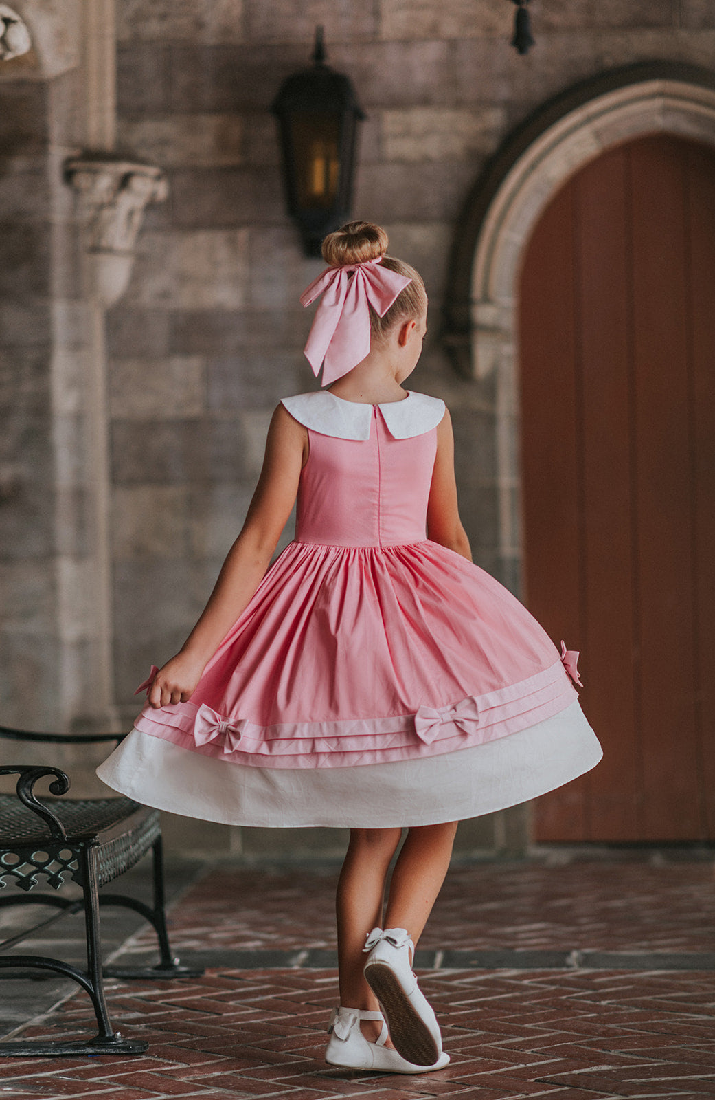 A young girl is wearing The Fairy Godmother Collection Cinderelly Pink Dress. The dress is woven cotton with a white collar and pink bow. The full skirt has bows at the bottom with a white hem. 