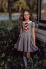 A young girl is wearing a grey Wizard Girl Twirl Dress with a striped yellow and brick red tie. The twirl dress has a white collar, short puff sleeves and a tie in the back at the waist. 