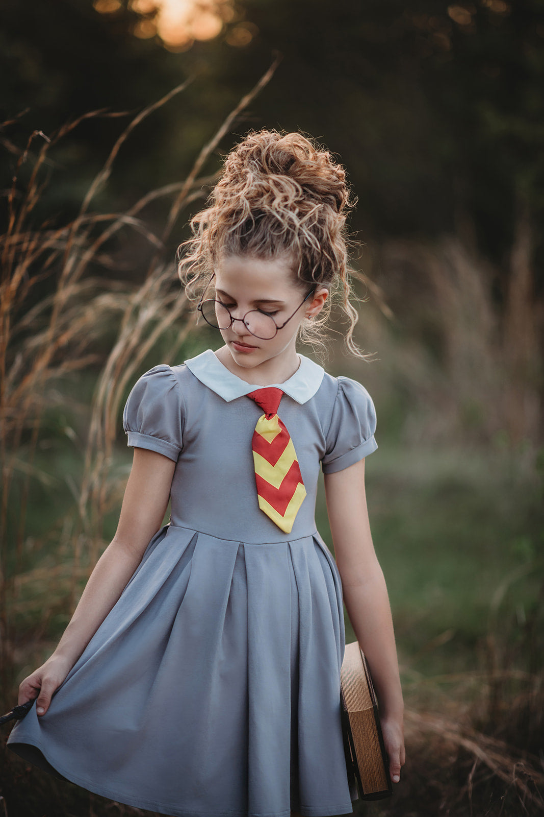 A young girl is wearing a grey Wizard Girl Twirl Dress with a striped yellow and brick red tie. The twirl dress has a white collar, short puff sleeves and a tie in the back at the waist. 