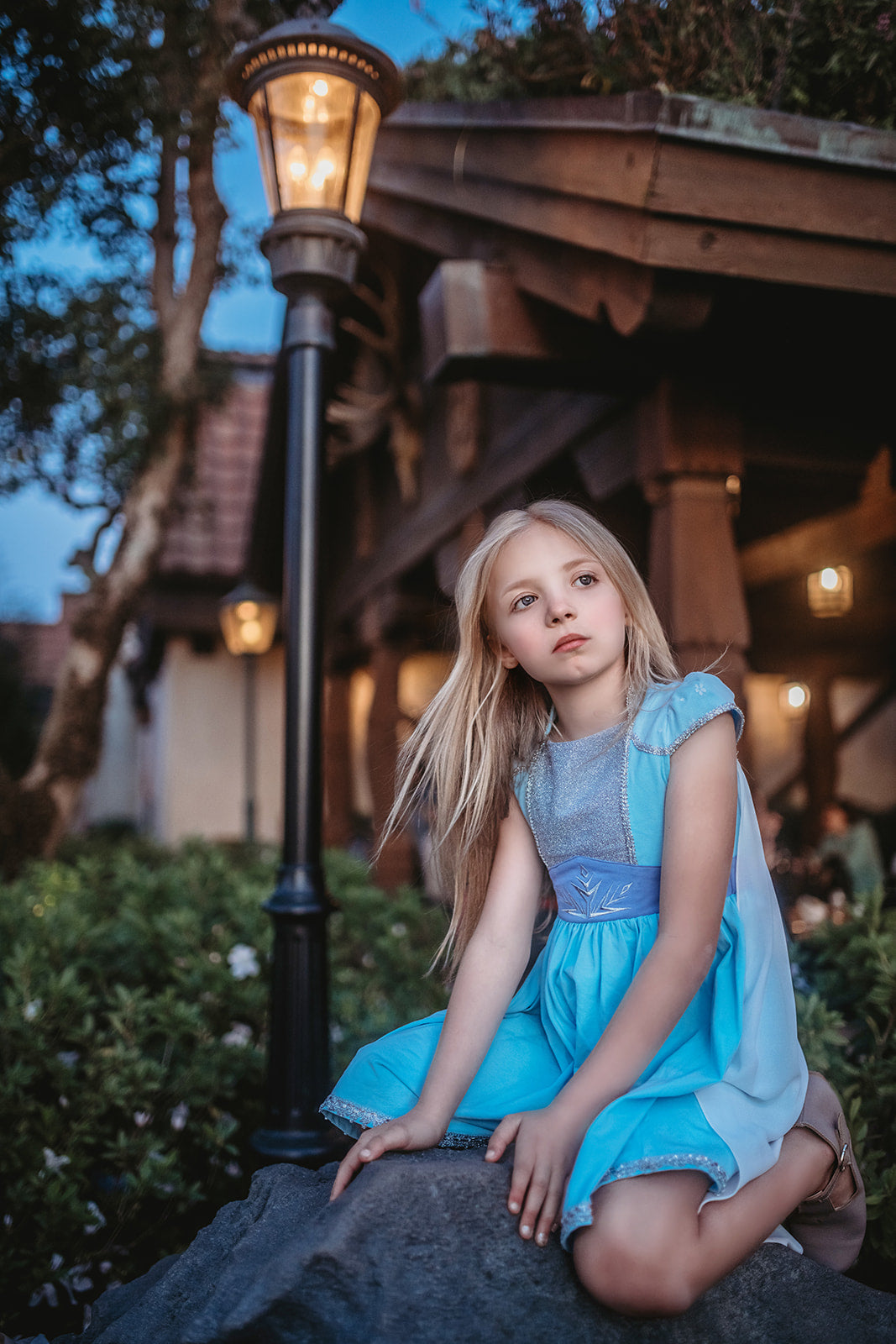 A young girl in an Ice Queen Dress and Cape Set, with a sheer chiffon removable cape. The light blue cap sleeves have white lace over them and silver racking on the bottom.  There is a silver sparkle panel on the chest and a silver embroidered snowflake on the darker blue fabric on the chest. Silver trim lines the bottom of the light blue twirl dress. 