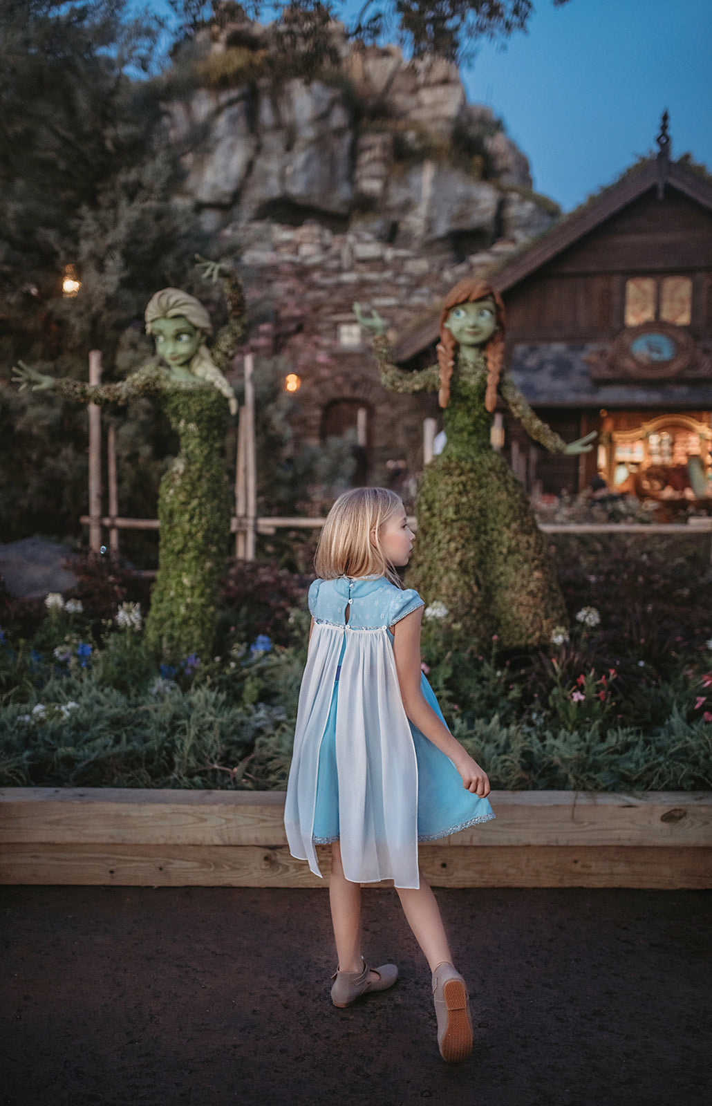 A young girl in an Ice Queen Dress and Cape Set, with a sheer chiffon removable cape. The light blue cap sleeves have white lace over them and silver racking on the bottom.  There is a silver sparkle panel on the chest and a silver embroidered snowflake on the darker blue fabric on the chest. Silver trim lines the bottom of the light blue twirl dress. 