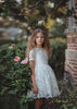 A young girl stands dressed in the Evangeline Frock in White. White tulle and lace dress features a built in petticoat for an extra full skirt and retro silhouette. The dress has cotton slip lining.