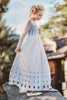 A young girl  is wearing the Queen of the Forest Maxi Dress adorned with blue diamond patterns along the bottom. The maxi dress also features a geometric design on the upper bodice and white mesh sleeves and neckline. The removable cape is fastened to the back with buttons. 