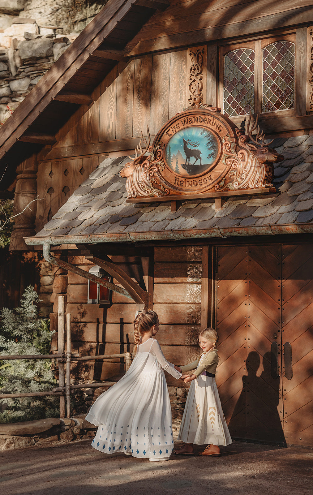 A young girl with blonde hair stands on a rock, wearing the Queen of the Forest Maxi Dress adorned with blue diamond patterns along the bottom. The maxi dress also features a geometric design on the upper part. She is looking downward and appears thoughtful, with a background of trees and buildings.