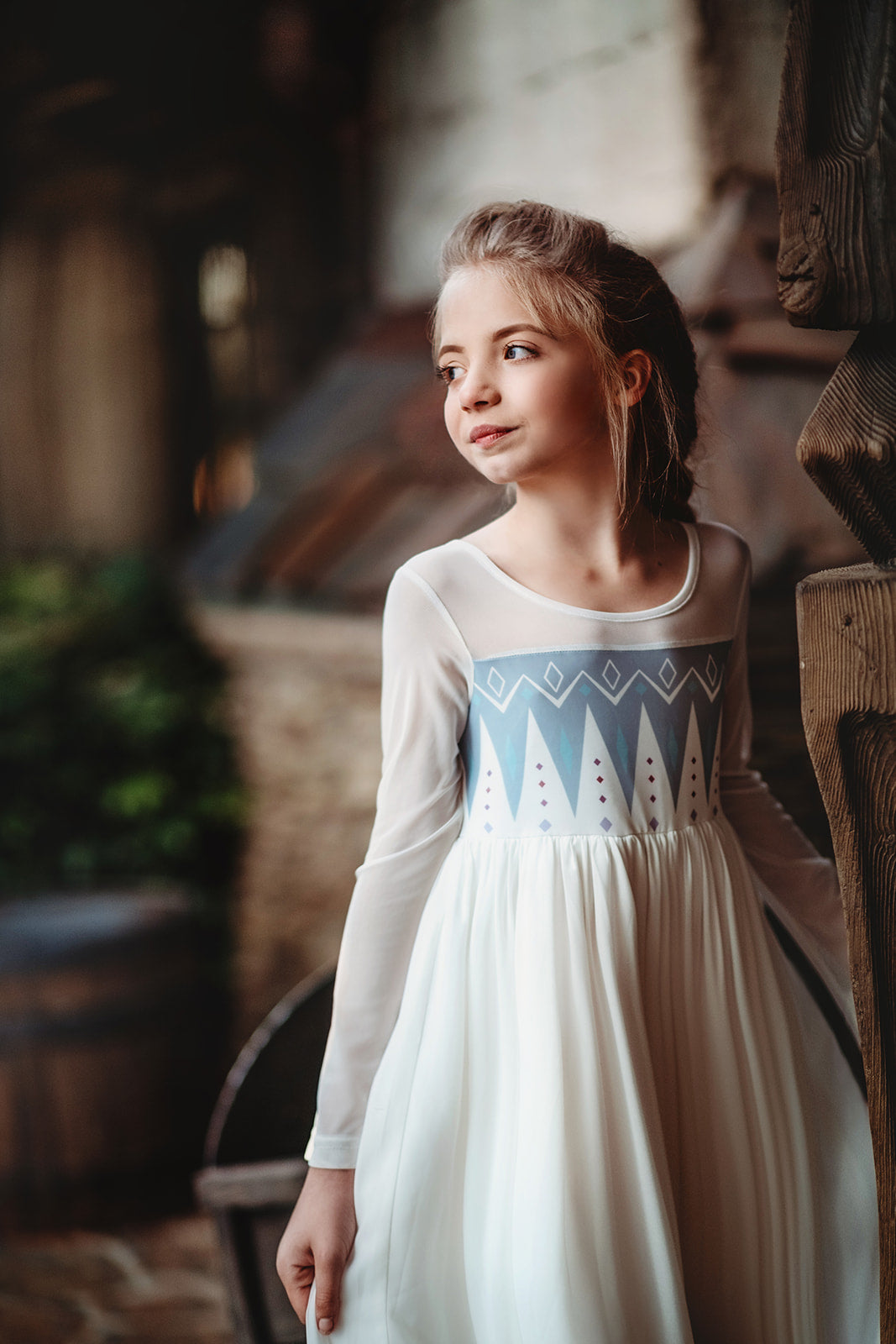 A young girl with blonde hair stands on a rock, wearing the Queen of the Forest Maxi Dress adorned with blue diamond patterns along the bottom. The maxi dress also features a geometric design on the upper part. She is looking downward and appears thoughtful, with a background of trees and buildings.