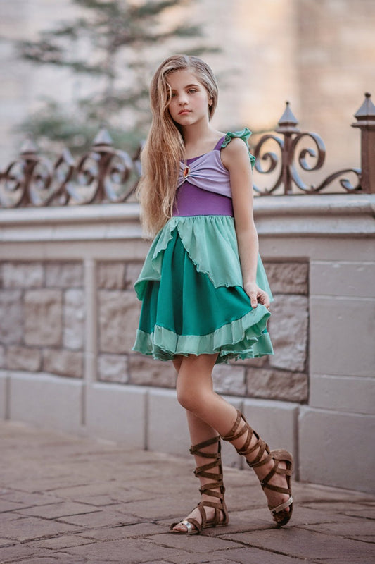 A young girl is wearing Our Original Mermaid Princess Twirl Dress in shades of green and purple. The sleeveless dress has green ruffle accent. The bodice has light and dark purple with a red ruby jewel. The twirl skirt is light and dark green with a ruffle at the hem. 