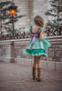 A young girl is wearing Our Original Mermaid Princess Twirl Dress in shades of green and purple. The sleeveless dress has green ruffle accent. The bodice has light and dark purple with a red ruby jewel. The twirl skirt is light and dark green with a ruffle at the hem. 