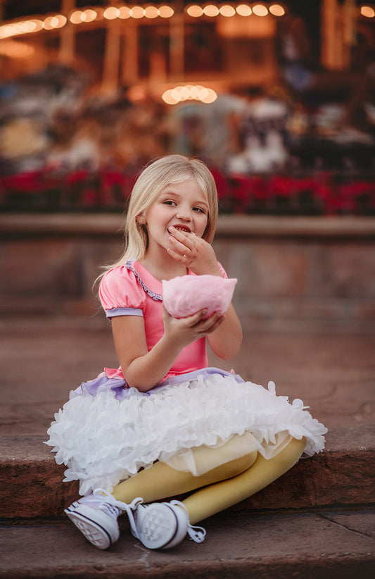 A young girl is wearing true pink and lavender bodice and white petal skirt dress.  The top has puff sleeves and ruffled collar.  The white skirt imitates feathers and is lined with tule and cotton to give a fuller appearance. 