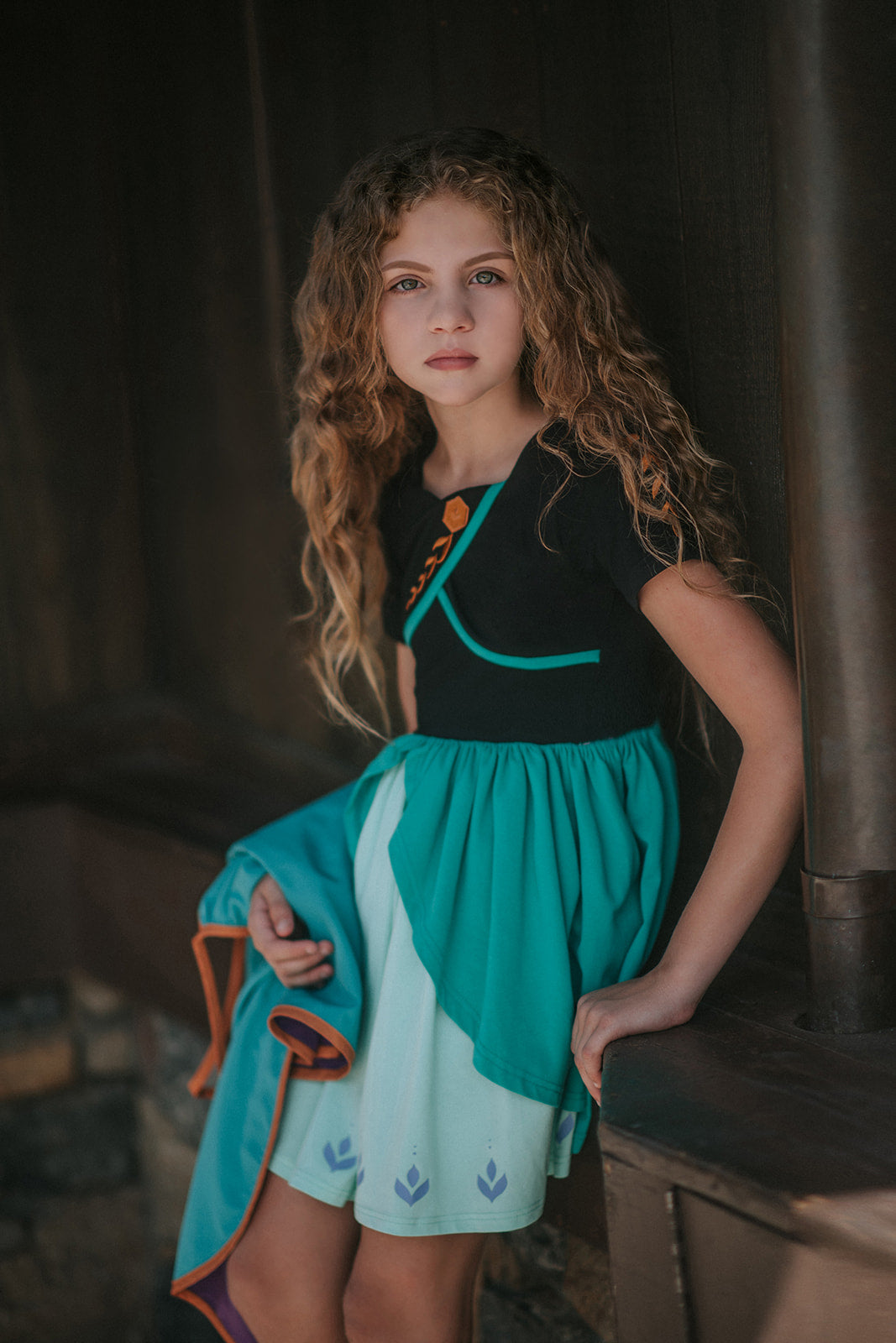 A young girl twirls in the vibrant Older Ice Princess Sister Set Style C dress, embellished with teal and purple accents. The removable cape is purple and teal with brown gold trim. The short sleeves feature a golden brown design.