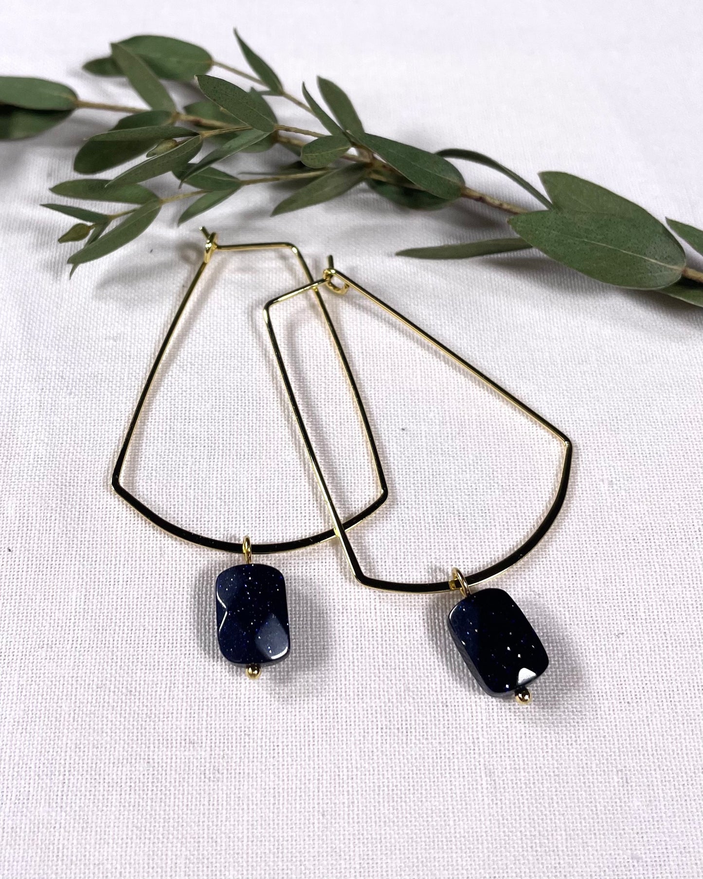 Golden hoops rectangle shape hoops with a dark purple blue with silver specks faceted stone that dangles from the bottom. 