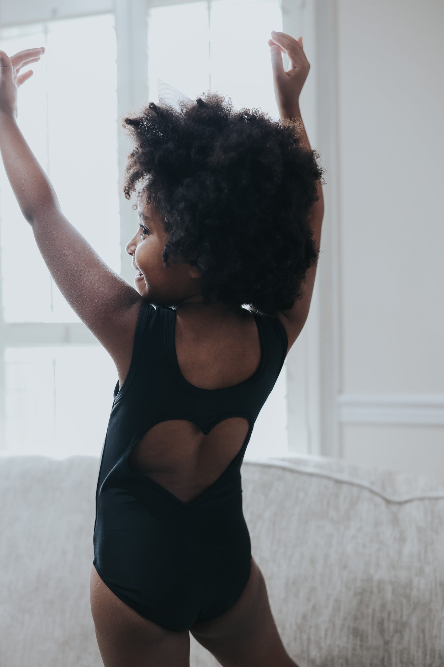 A young girl is wearing the Love Bug Leotard, a black gymnastics outfit with an elegant heart cutout back. 