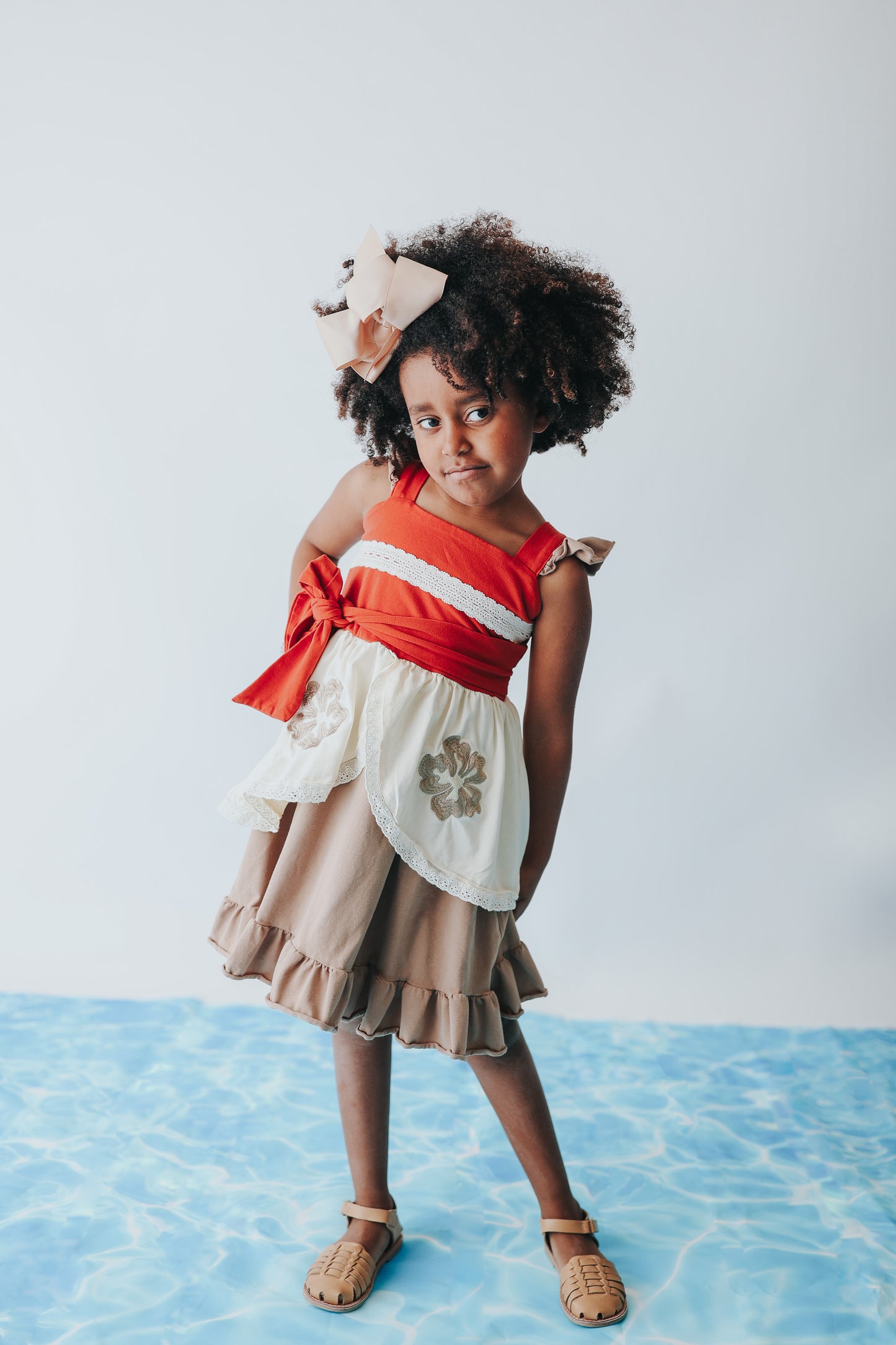 A young girl is wearing the Polynesian Princess Twirl Dress, which features beige and cream fabric accented by a red waistband and decorative floral embroidery.  There is a lace accent on the chest and ruffles on the top of the straps.  There is a ruffle on the hem and a tie on the side of the dress. Perfect for an adventurous princess. 