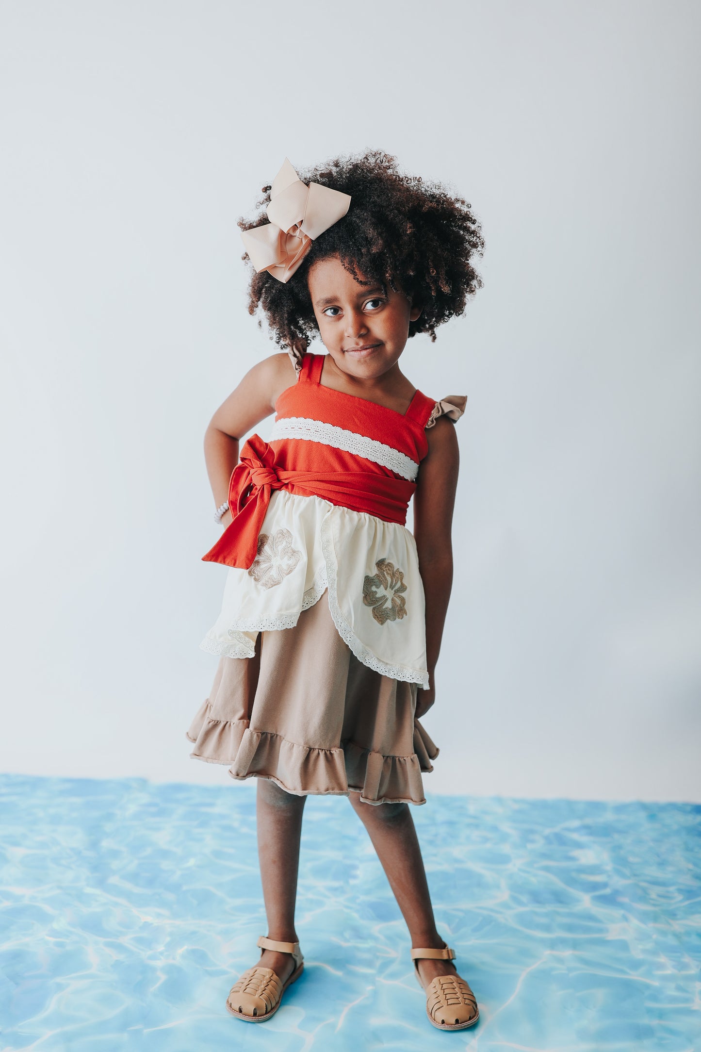 A young girl is wearing the Polynesian Princess Twirl Dress, which features beige and cream fabric accented by a red waistband and decorative floral embroidery.  There is a lace accent on the chest and ruffles on the top of the straps.  There is a ruffle on the hem and a tie on the side of the dress. Perfect for an adventurous princess. 