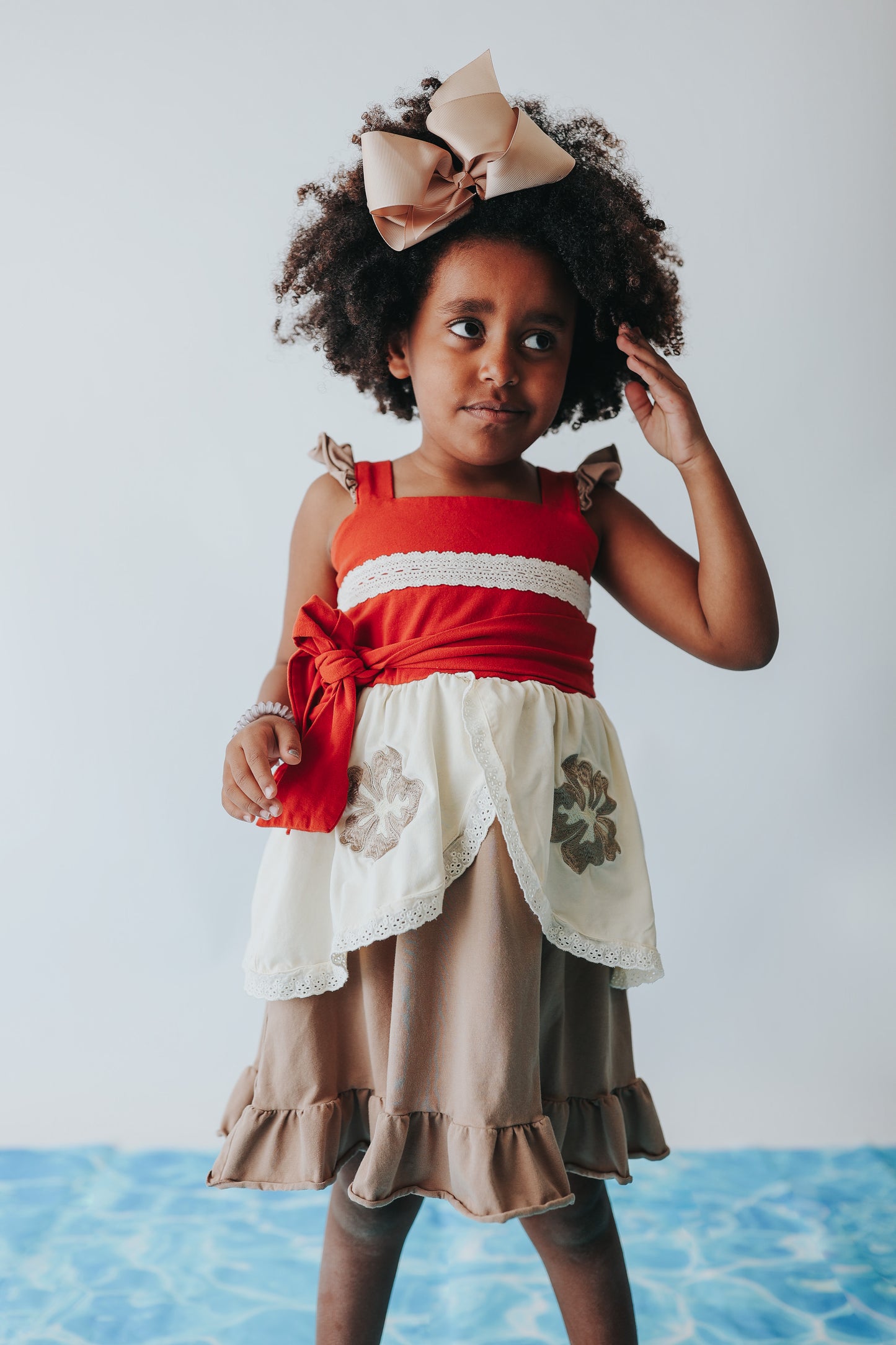 A young girl is wearing the Polynesian Princess Twirl Dress, which features beige and cream fabric accented by a red waistband and decorative floral embroidery.  There is a lace accent on the chest and ruffles on the top of the straps.  There is a ruffle on the hem and a tie on the side of the dress. Perfect for an adventurous princess. 