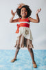 A young girl with curly hair stands barefoot on a grassy area, holding a carved wooden paddle. She is wearing the "Our Original Polynesian Princess Twirl Dress," which features beige and white fabric accented by a red waistband and decorative floral embroidery, evoking the essence of a Polynesian Princess, with greenery and a tree trunk in the background.