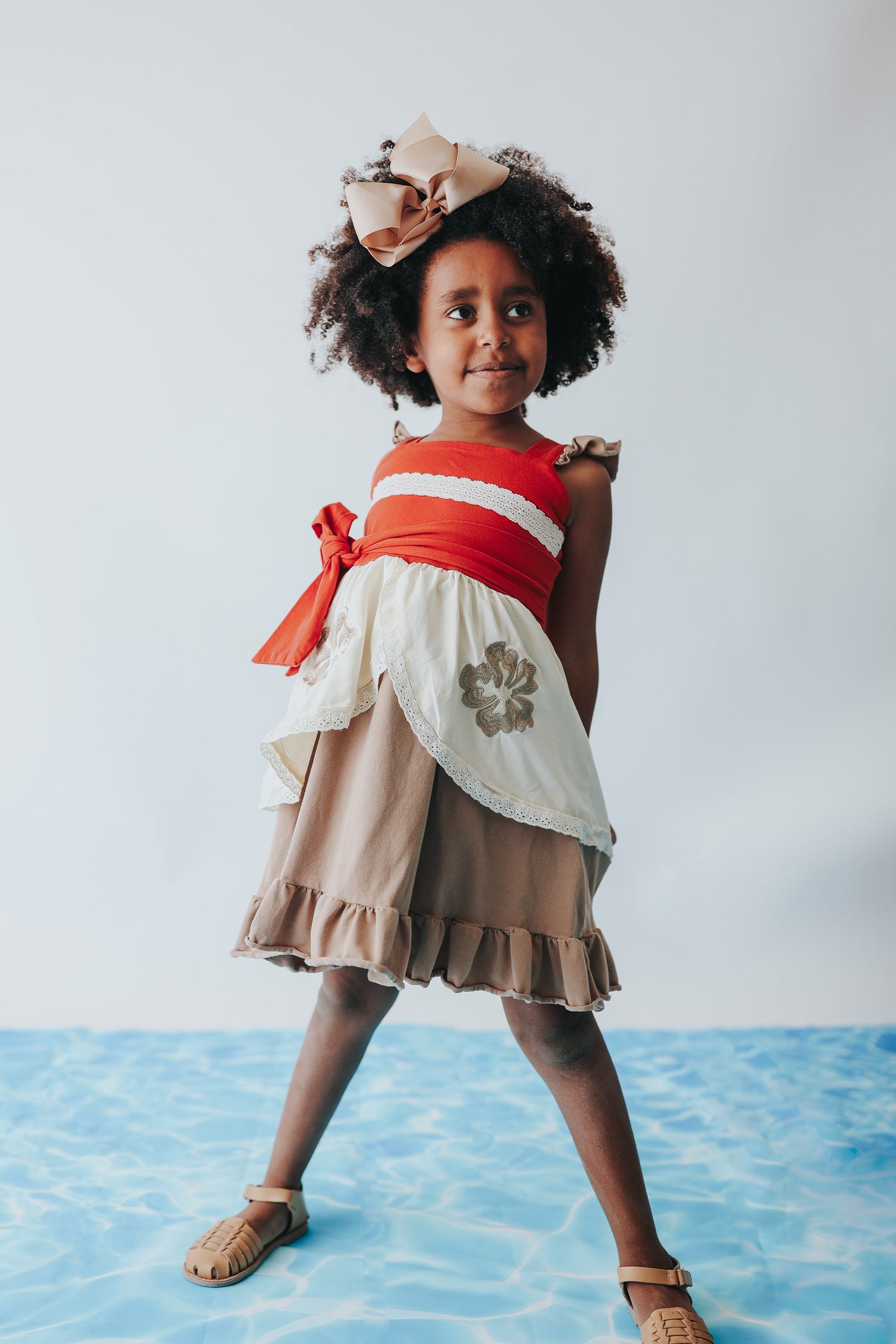 A young girl is wearing the Polynesian Princess Twirl Dress, which features beige and cream fabric accented by a red waistband and decorative floral embroidery.  There is a lace accent on the chest and ruffles on the top of the straps.  There is a ruffle on the hem and a tie on the side of the dress. Perfect for an adventurous princess. 