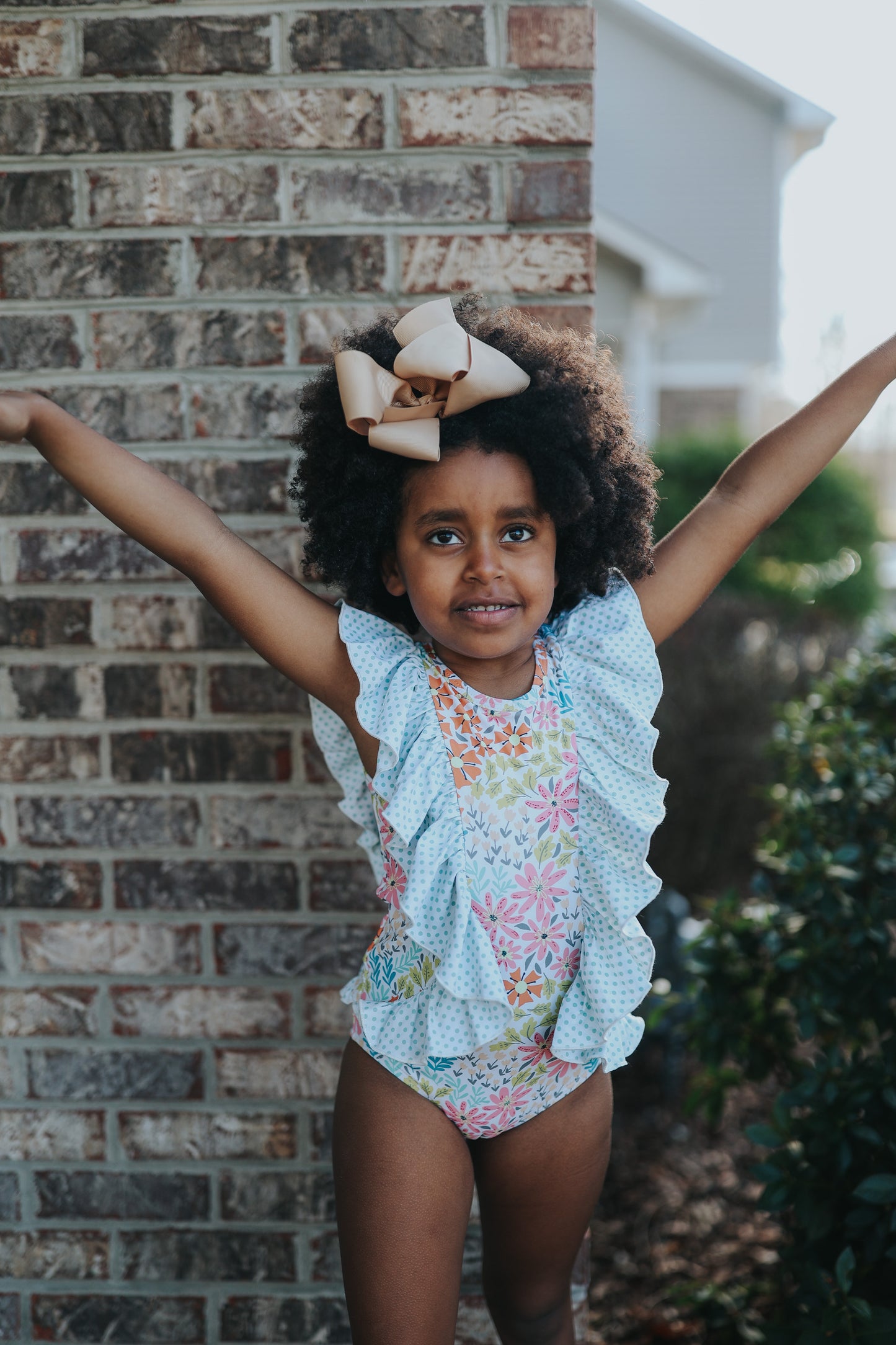 A young girl smiles brightly, wearing the Nora Leotard in Wildflower, adorned with a beautiful floral pattern and ruffles. The ruffles are light blue polka dot.  The wildflower body has pink, orange, blue green and light yellow. 