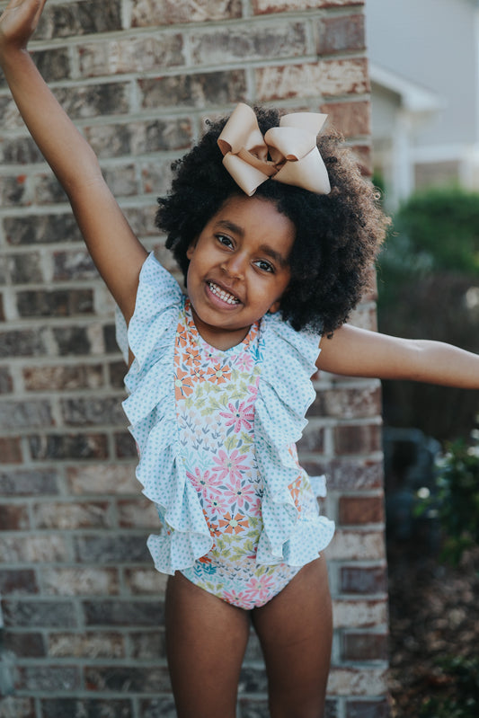 A young girl with curly hair smiles brightly, wearing the Nora Leotard in Wildflower, adorned with a beautiful floral pattern and ruffles. She has a large beige bow in her hair and stands against a brick wall with arms extended playfully.