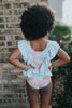 A young girl smiles brightly, wearing the Nora Leotard in Wildflower, adorned with a beautiful floral pattern and ruffles. The ruffles are light blue polka dot.  The wildflower body has pink, orange, blue green and light yellow. 