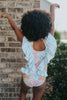 A young girl smiles brightly, wearing the Nora Leotard in Wildflower, adorned with a beautiful floral pattern and ruffles. The ruffles are light blue polka dot.  The wildflower body has pink, orange, blue green and light yellow. 