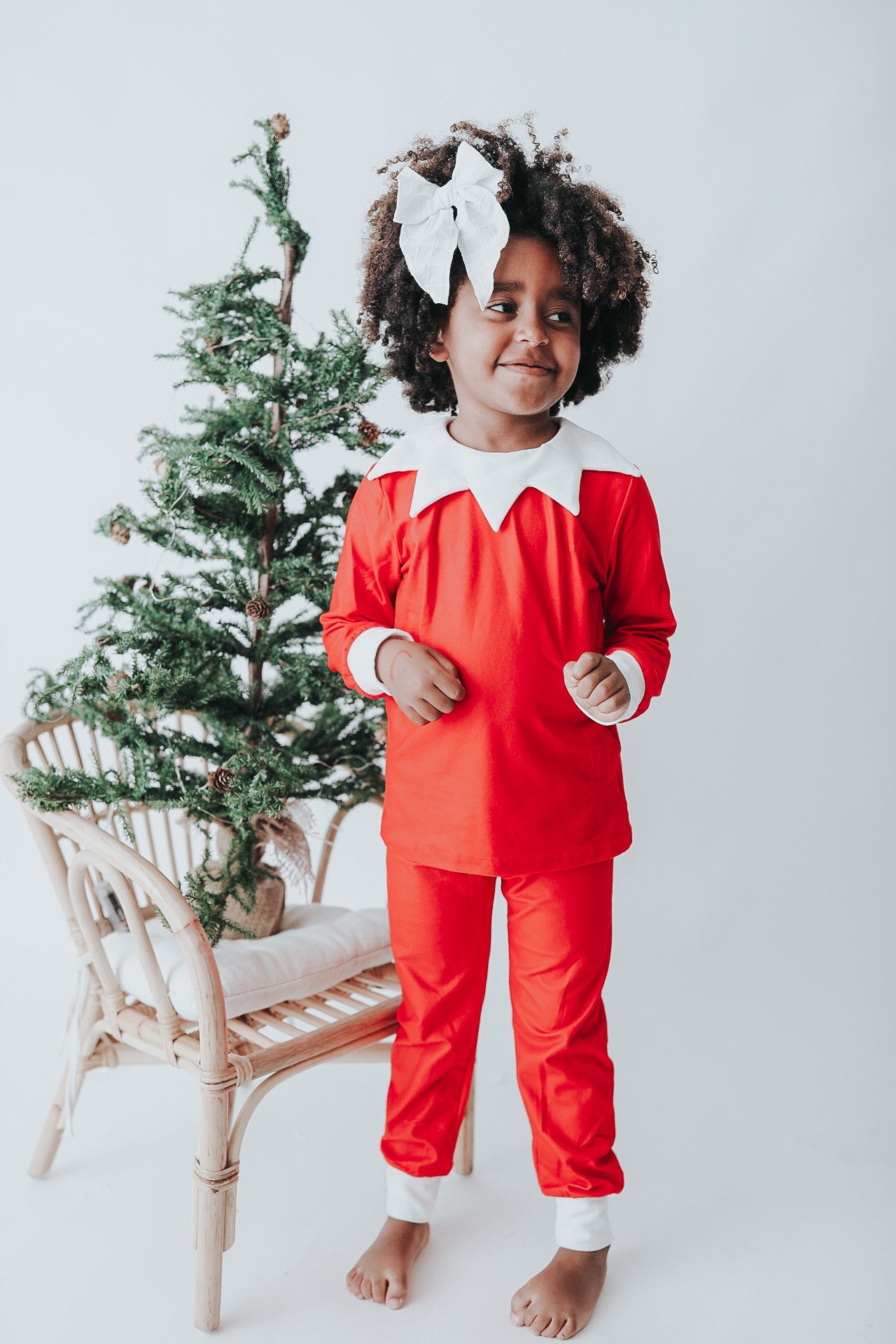 A young child dressed in Unisex Elf Pajamas with a red pointy hat. The perfect Christmas photo outfit. The fabric is bright red and buttery soft.  The top is long sleeved with white cuffs and a white collar that has large triangle points similar to a clown. Long pants with white cuffs. 