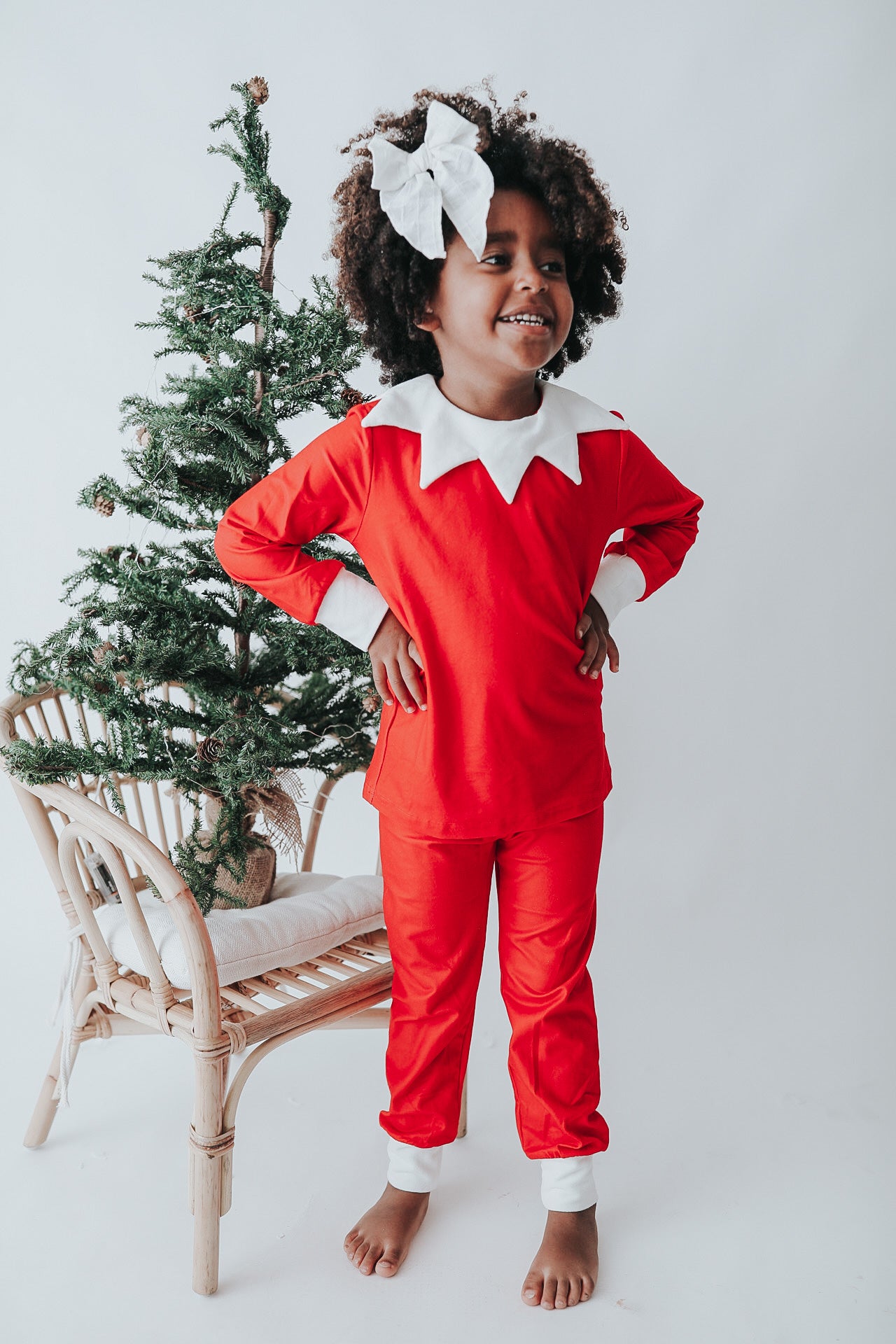 A young child dressed in Unisex Elf Pajamas with a red pointy hat. The perfect Christmas photo outfit. The fabric is bright red and buttery soft.  The top is long sleeved with white cuffs and a white collar that has large triangle points similar to a clown. Long pants with white cuffs. 