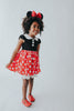 A young girl is wearing the Miss Mouse Twirl Dress, which features a black bodice, puff sleeves and a white collar.  Three white buttons decorate the center. The skirt is red with white polka dots and white eyelet lace trim.