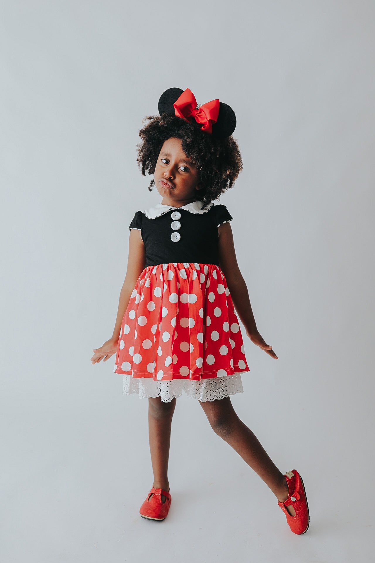 A young girl is wearing the Miss Mouse Twirl Dress, which features a black bodice, puff sleeves and a white collar.  Three white buttons decorate the center. The skirt is red with white polka dots and white eyelet lace trim.