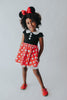 A young girl is wearing the Miss Mouse Twirl Dress, which features a black bodice, puff sleeves and a white collar.  Three white buttons decorate the center. The skirt is red with white polka dots and white eyelet lace trim.