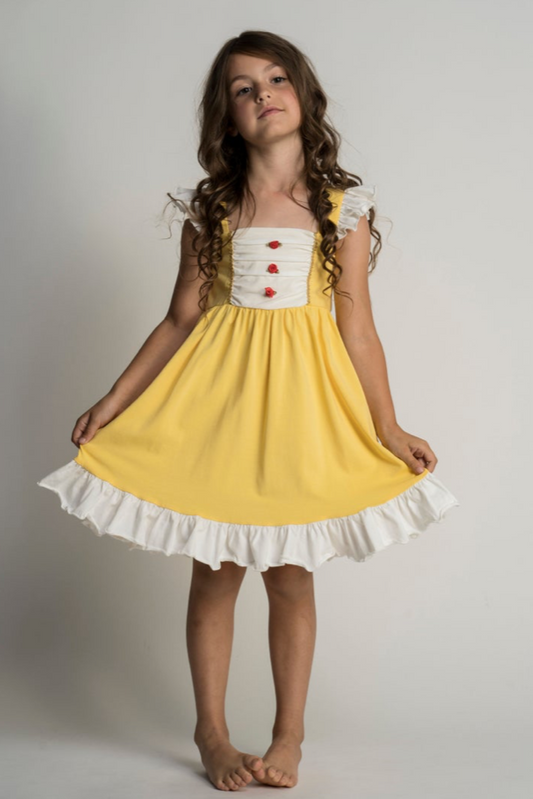 A young girl with long, wavy hair stands barefoot against a plain backdrop, wearing the Our Original Red Rose Twirl Dress from the Princess Twirl Collection. The sleeveless yellow dress features white ruffled trim and red buttons down the front. She holds the sides of her dress out slightly, creating a gentle arc.