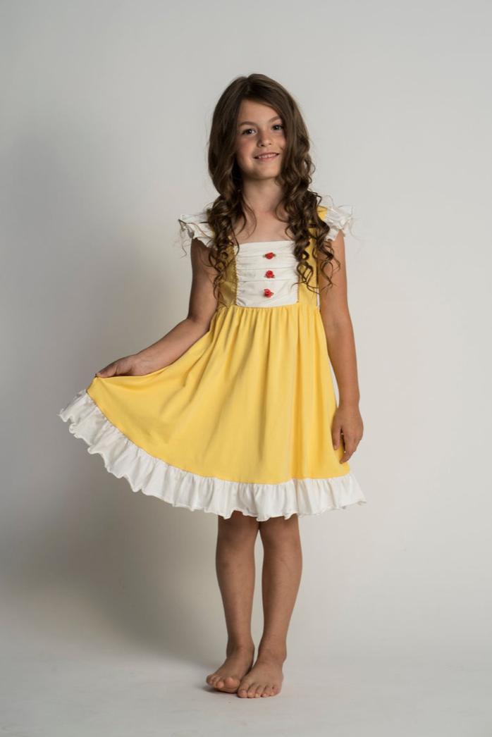 A young girl with long, wavy hair stands barefoot against a plain backdrop, wearing the Our Original Red Rose Twirl Dress from the Princess Twirl Collection. The sleeveless yellow dress features white ruffled trim and red roses down the front. She holds the sides of her dress out slightly, creating a gentle arc.