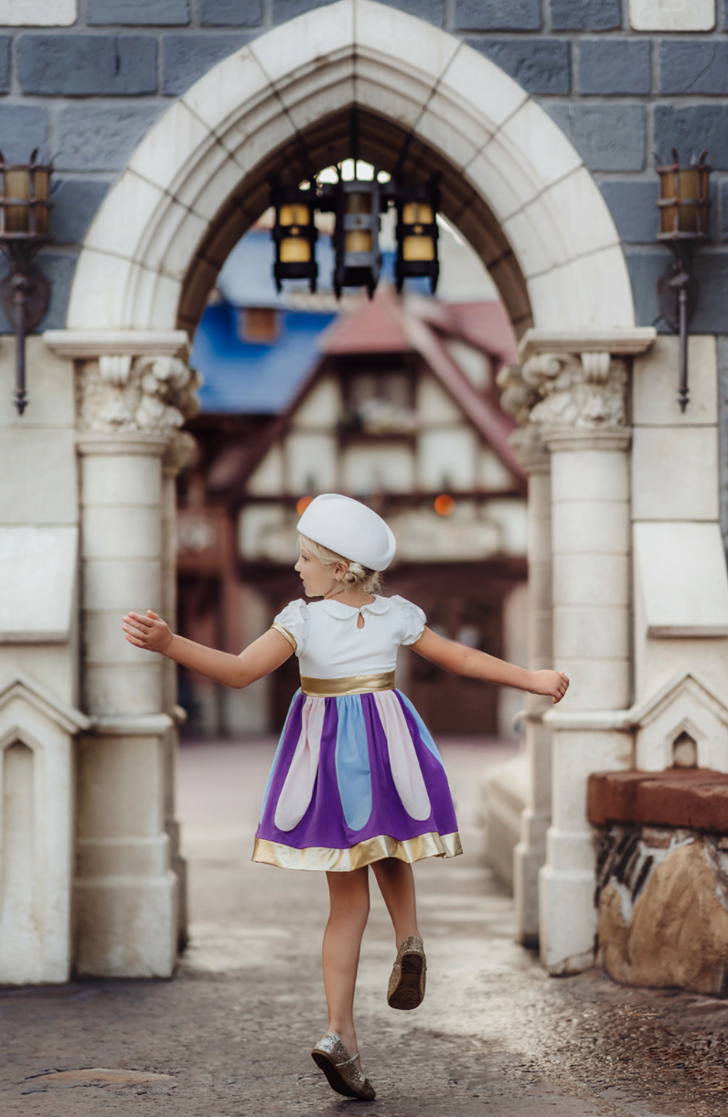 A young girl is twirling in a cotton dress. The bodice has a peter pan collar and puff sleeves with gold cuffs. There is a band of gold at the waist. The skirt is purple with light pink and light blue panels. A metallic gold band lines the hem. 