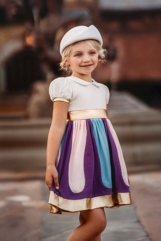 A young girl with blonde hair stands outdoors wearing a white hat and a colorful dress from the "I'm a Little Teacup Twirl" collection, featuring purple, pink, blue, and gold accents. Made from soft cotton jersey knit fabric, she smiles happily against a slightly blurred scenic background.