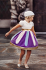 A young girl is twirling in a cotton dress. The bodice has a peter pan collar and puff sleeves with gold cuffs. There is a band of gold at the waist. The skirt is purple with light pink and light blue panels. A metallic gold band lines the hem. 